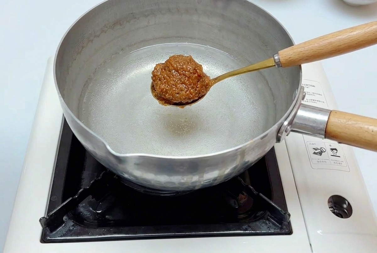 Ein goldener Löffel mit der Miso-Mischung wird in einen Edelstahltopf mit Wasser auf dem Herd getaucht.