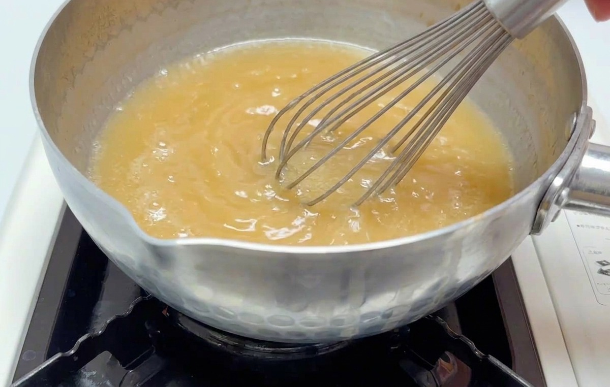 Eine Hand verquirlt mit einem Schneebesen eine trübe, hellbraune Miso-Brühe in einem kleinen Topf auf einer eingeschalteten Herdplatte.