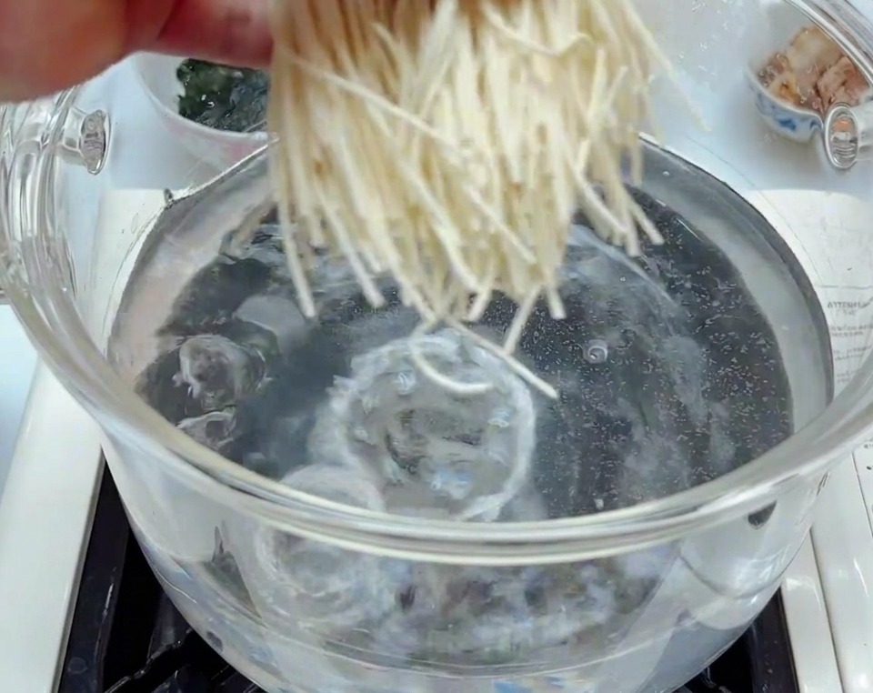 Frische, weiße Ramen-Nudeln werden in einen durchsichtigen Glastopf mit sprudelnd kochendem Wasser gegeben.