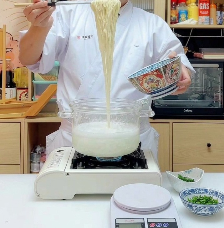 Eine Portion langer, gelber Ramen-Nudeln wird mit Stäbchen hoch aus einem dampfenden Topf gehoben.