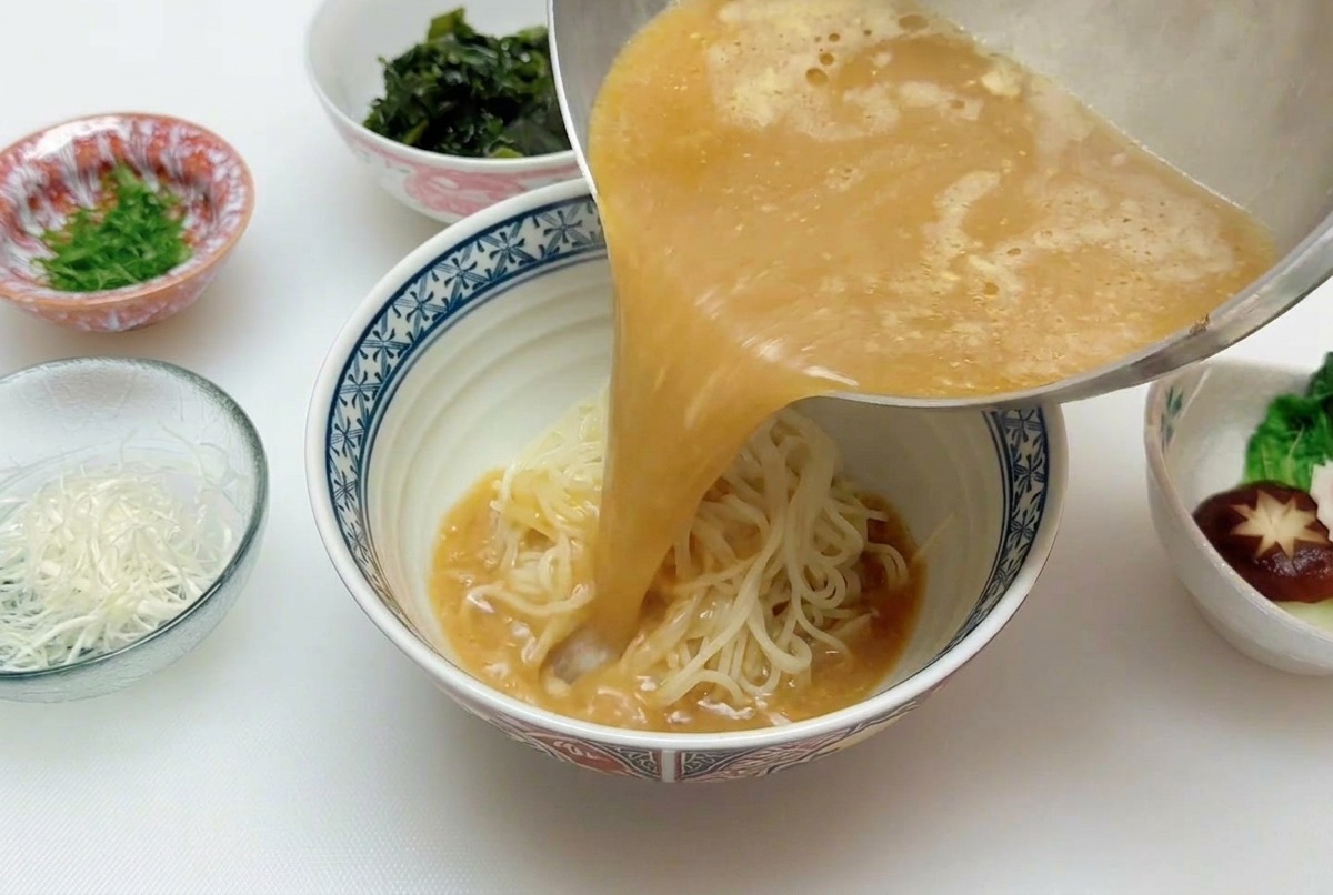Ein stetiger Strahl heißer, goldbrauner Miso-Brühe wird aus einem kleinen Topf über einen Berg Ramen-Nudeln in einer Schüssel gegossen.