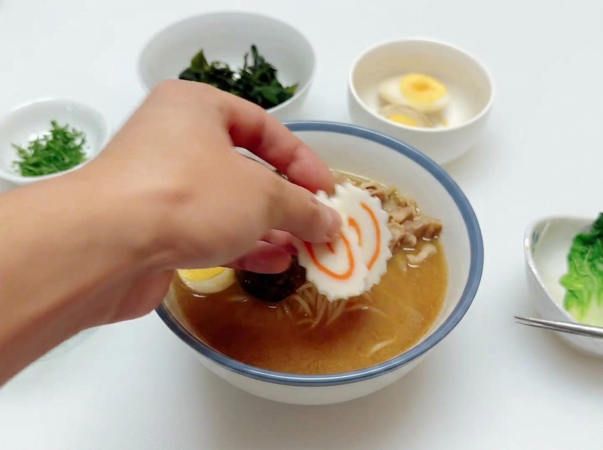 Eine Hand legt eine Scheibe rosa-weiß gestrudelter Narutomaki auf eine Ramen-Schüssel, die bereits Rindfleisch und Ei enthält.