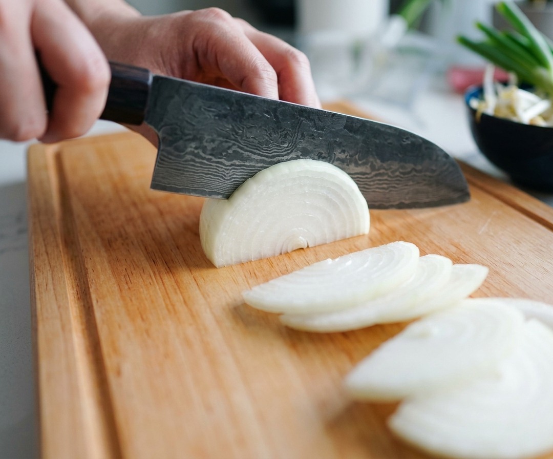 Eine person schneidet eine weiße Zwiebel mit einem Kochmesser in feine Halbmonde.