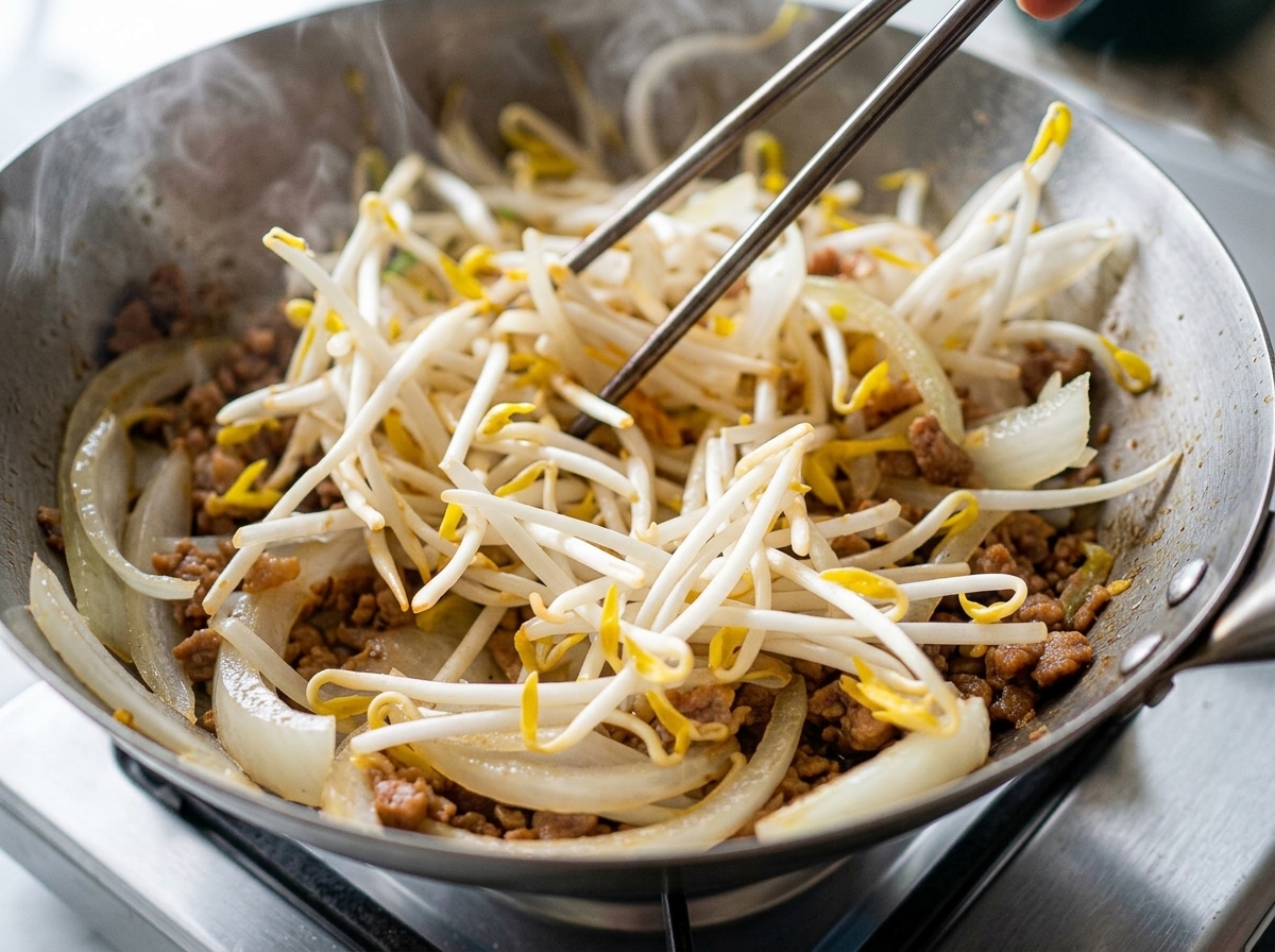 Eine große Portion weiße Sojasprossen wird zu dem Fleisch und den Zwiebeln in die Pfanne gegeben.