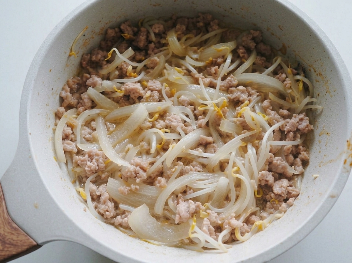 Eine Mischung aus gedünsteten Zwiebeln, Sprossen und Hackfleisch wird in einer hellen Pfanne gewendet.
