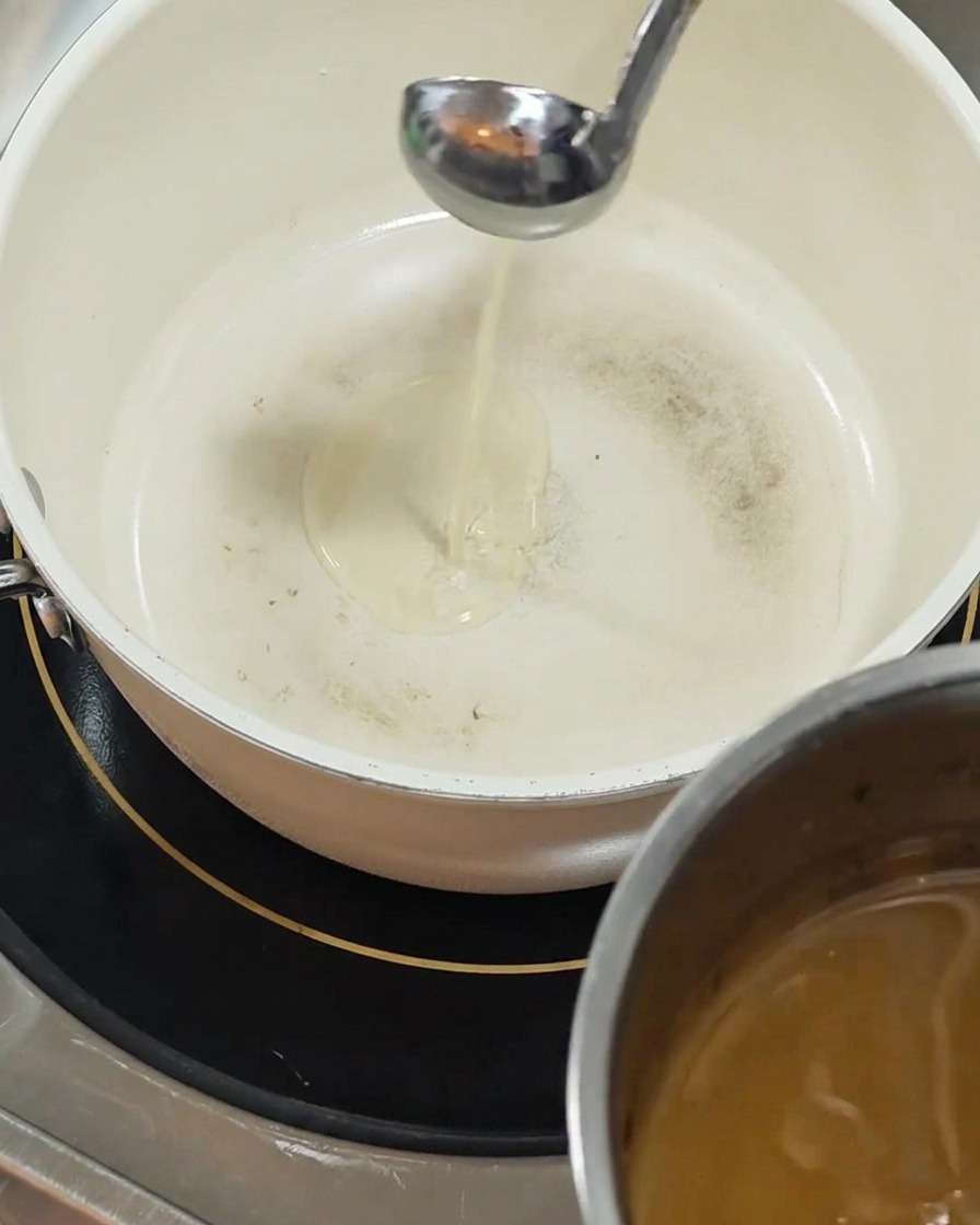 Eine Schöpfkelle aus Silber gießt klares, gelbes Öl in einen weißen, keramikbeschichteten Topf auf einer schwarzen Induktionskochplatte.