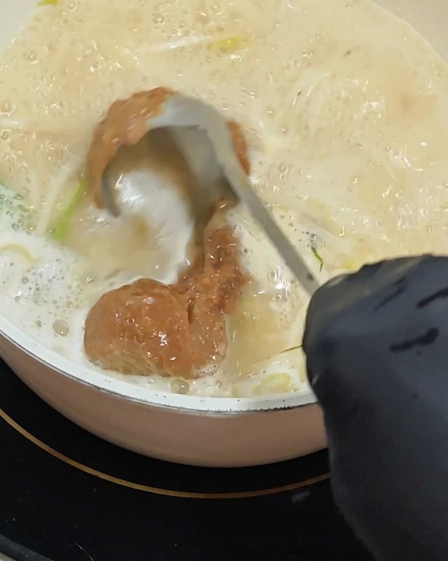 Eine Hand mit Handschuh rührt mit einer Schöpfkelle eine dicke Portion rötlich-braune Misopaste in eine leicht köchelnde, cremige Suppenbasis ein.