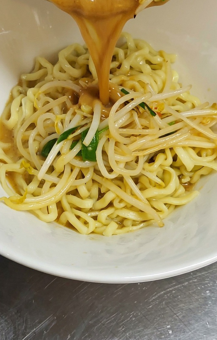 Eine kräftige, braune Miso-Brühe samt angebratenen Sojasprossen wird aus einem Topf in eine weiße Schüssel gegossen, die mit gewellten gelben Ramen-Nudeln gefüllt ist.