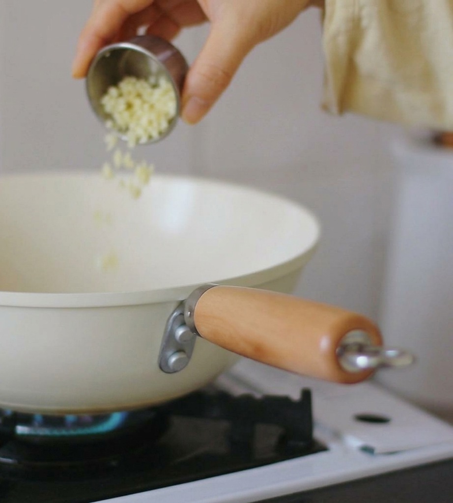 Eine Hand gießt fein gehackten Knoblauch aus einer kleinen Edelstahlschüssel in eine cremefarbene Pfanne auf einem Gasherd mit blauer Flamme.