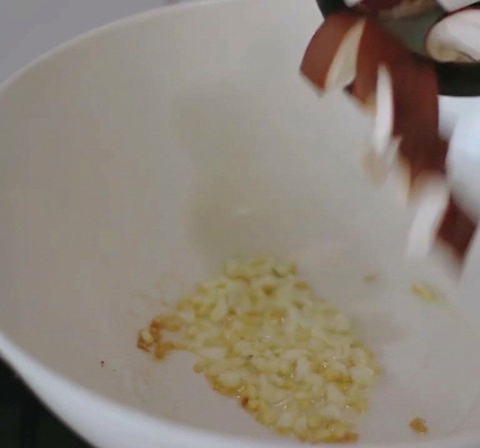 In einer weißen Pfanne auf dem Herd befindet sich angebratener Knoblauch, zu dem in Scheiben geschnittene Shiitake-Pilze von oben hinzugefügt werden.