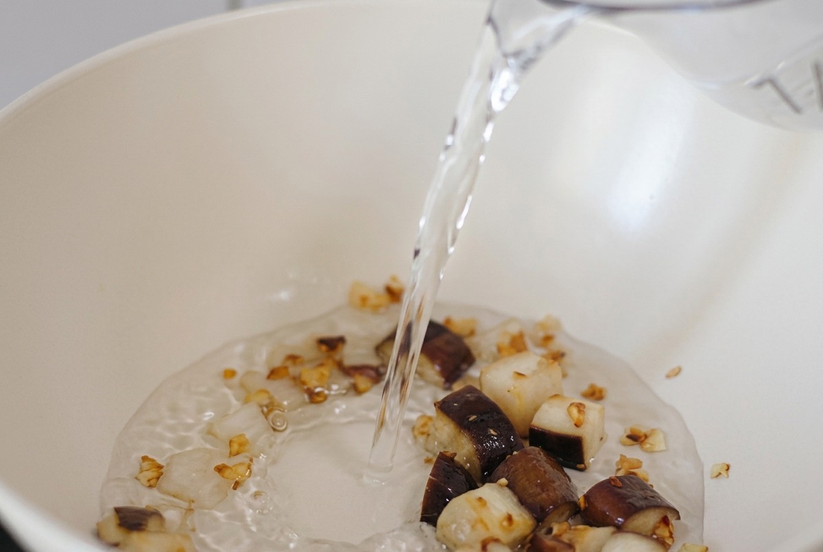 Wasser wird aus einer Kanne in eine weiße Pfanne mit angebratenen Pilz- und Knoblauchstücken gegossen.