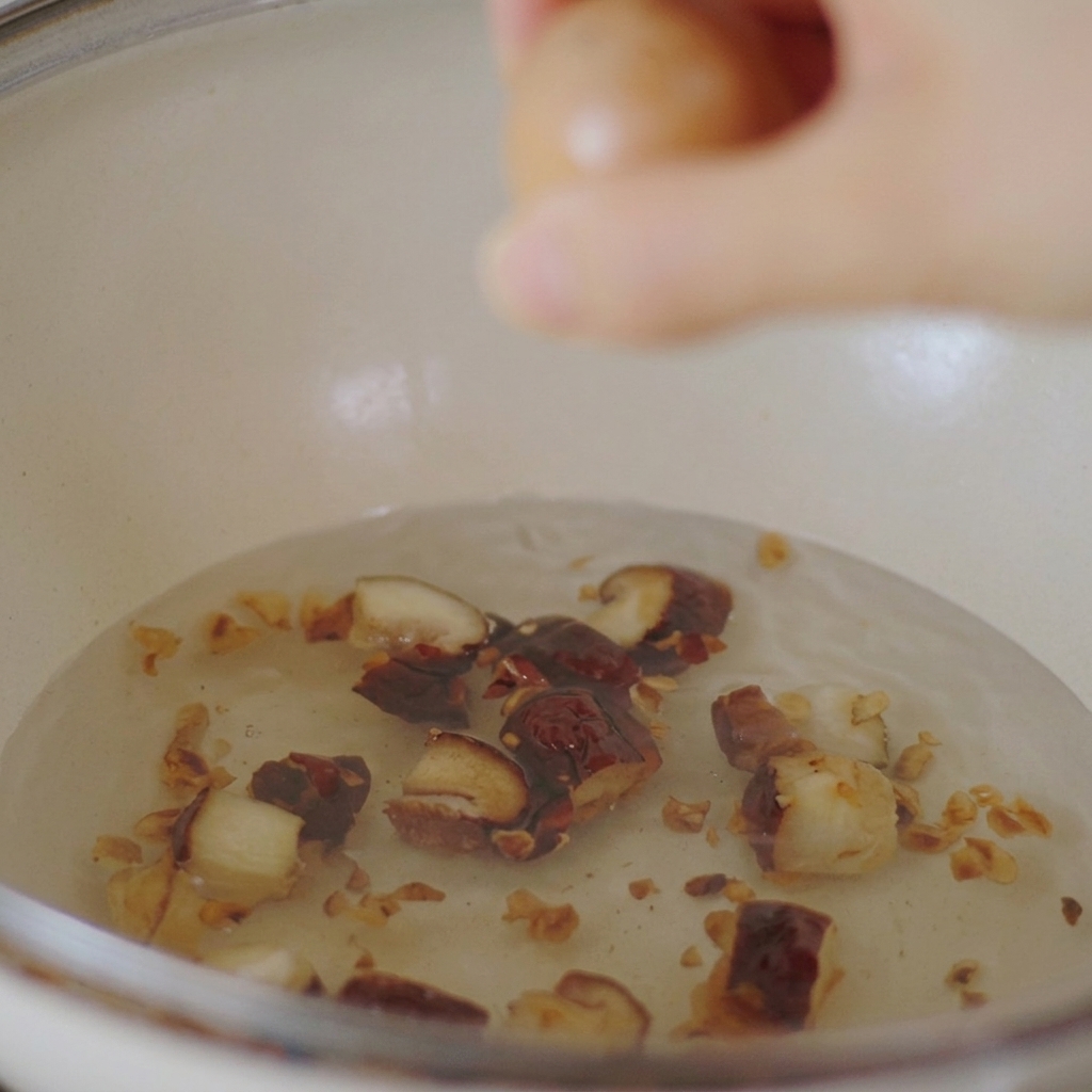 Eine Hand lässt ein braunes Hühnerei vorsichtig in einen weißen Topf mit Wasser und geschnittenen Shiitake-Pilzen gleiten.