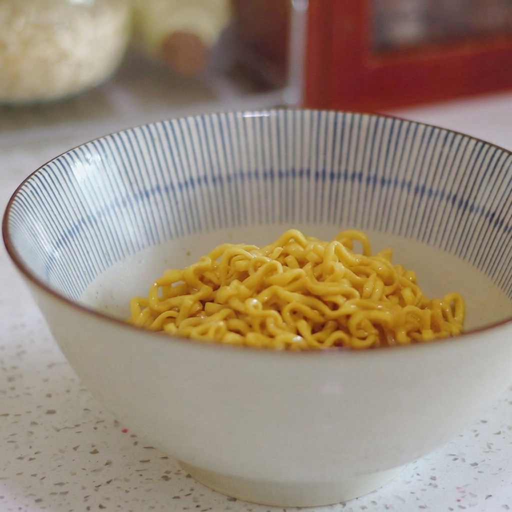Gelbe, lockige Ramen-Nudeln liegen am Boden einer blau-weiß gestreiften Keramikschüssel.