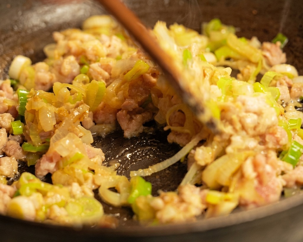 Ein Holzpfannenwender rührt gebräuntes Schweinehackfleisch und Frühlingszwiebeln in einer heißen, dampfenden Pfanne um.