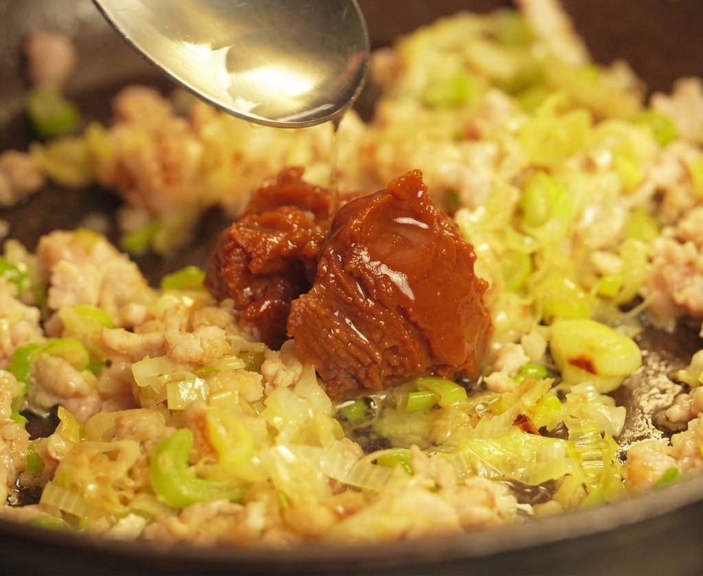 Ein silberner Löffel gibt einen großen Klecks dunkle Misopaste auf das angebratene Schweinehackfleisch und die Zwiebeln in der Pfanne.