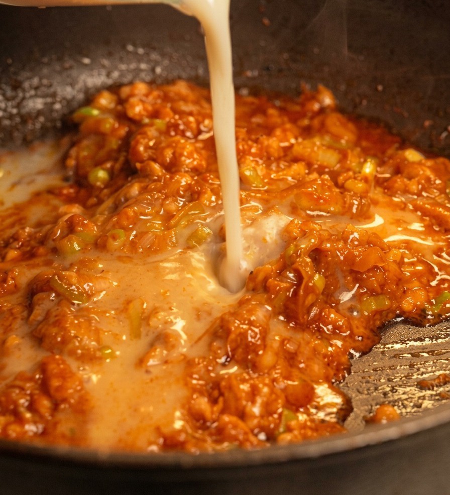 Ein Strahl cremeweißer Sojamilch wird in eine Pfanne mit der rötlichen, herzhaften Fleisch-Miso-Mischung gegossen.