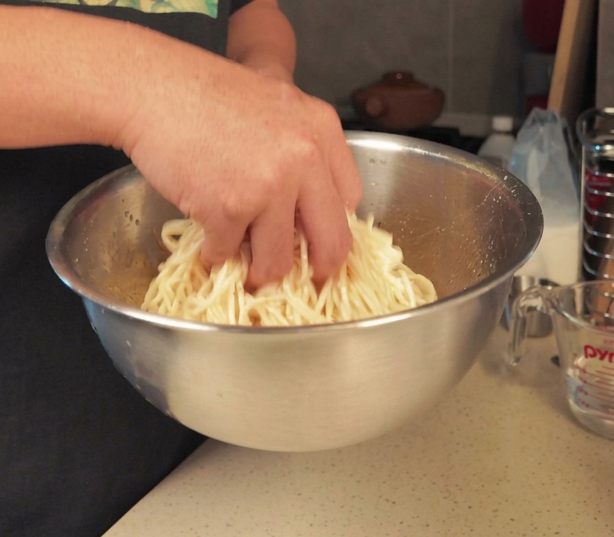 Eine Person vermengt eine Portion gekochte weiße Nudeln mit der Hand in einer großen Edelstahlschüssel, um sie mit Öl zu benetzen.