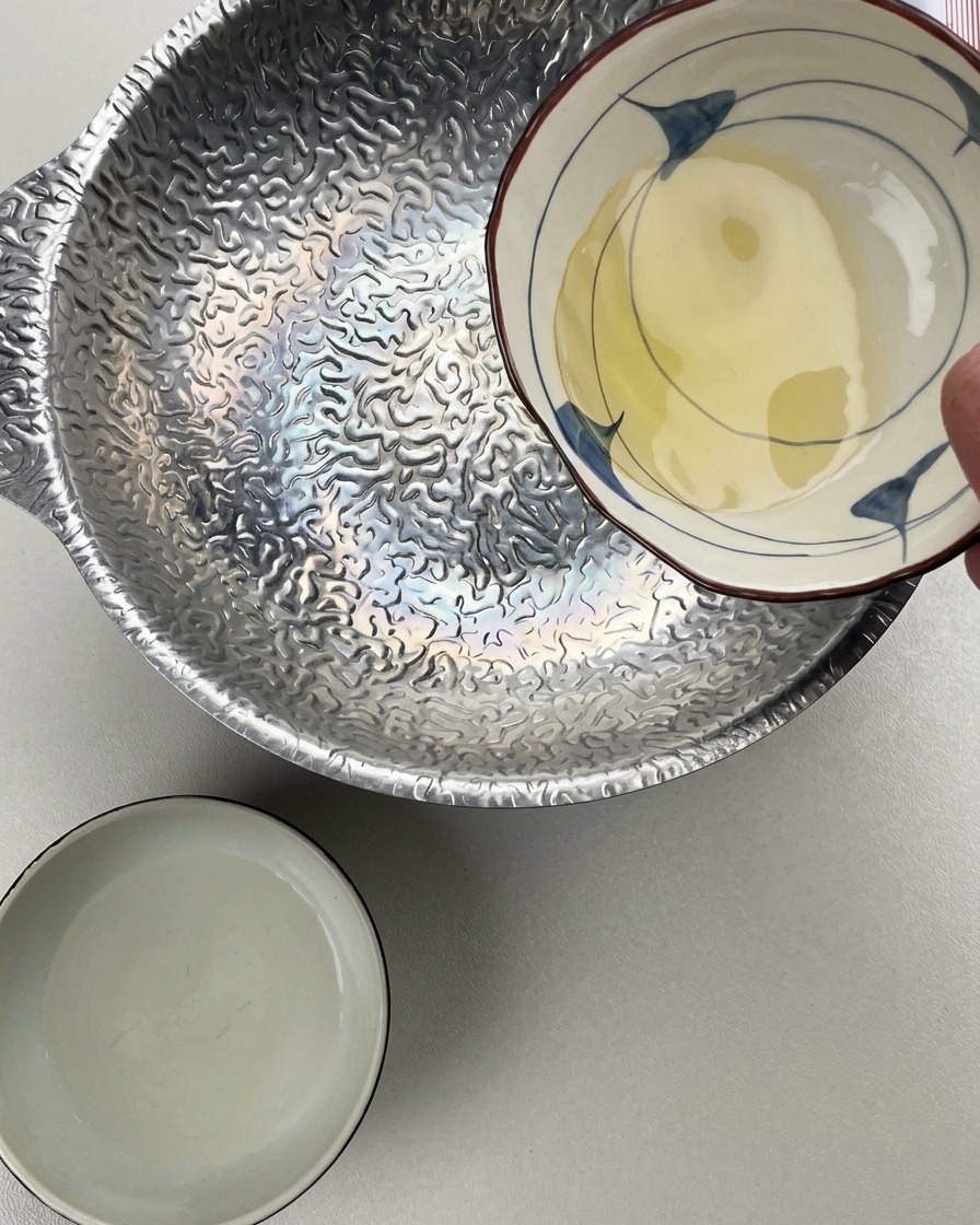 Eine Person gießt Mirin aus einer kleinen Keramikschale in einen silbernen, strukturierten Topf, während eine weitere Schale mit Sake bereitsteht.