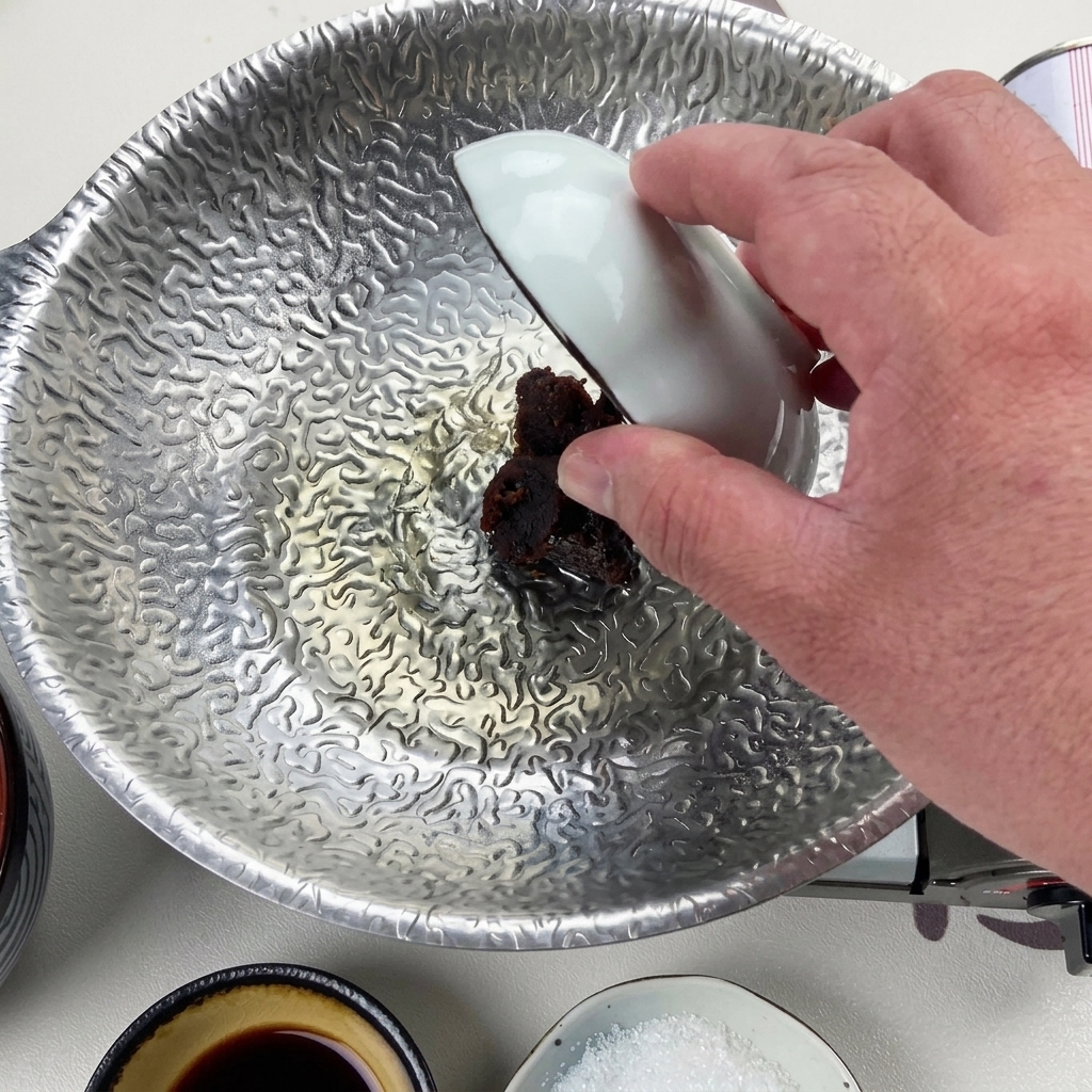 Eine Hand gibt mit einer kleinen weißen Schale eine kräftige Portion dunkle rote Misopaste in die reduzierte Flüssigkeit am Boden des silbernen Topfes.