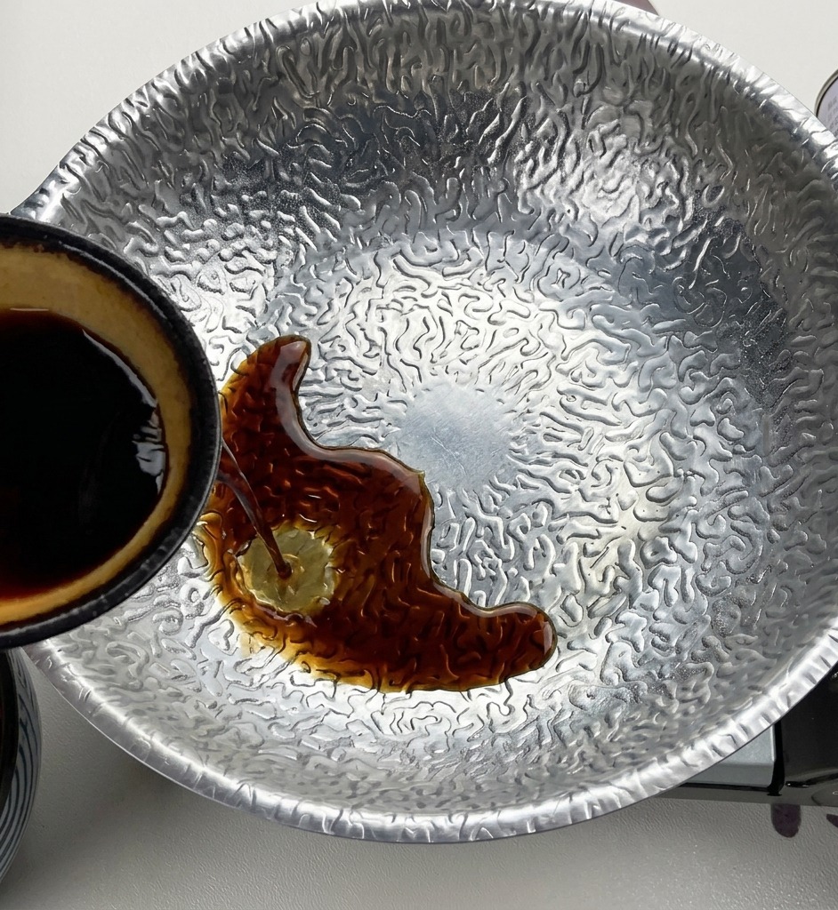Dunkle Sojasauce wird aus einer kleinen Schale mit gelber Innenseite in einen strukturierten Metalltopf mit Miso und Zucker gegossen.