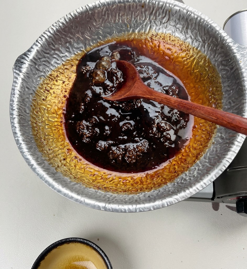 Ein Holzlöffel rührt eine dicke, dunkelbraune, köchelnde Sauce in einem gehämmerten, silberfarbenen Topf.