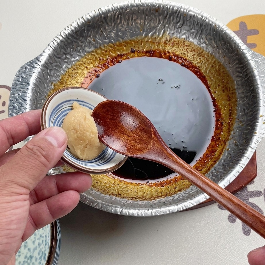 Ein Holzlöffel hält eine Portion helle weiße Misopaste über eine dunkle, eingekochte Sauce in einem Metalltopf.