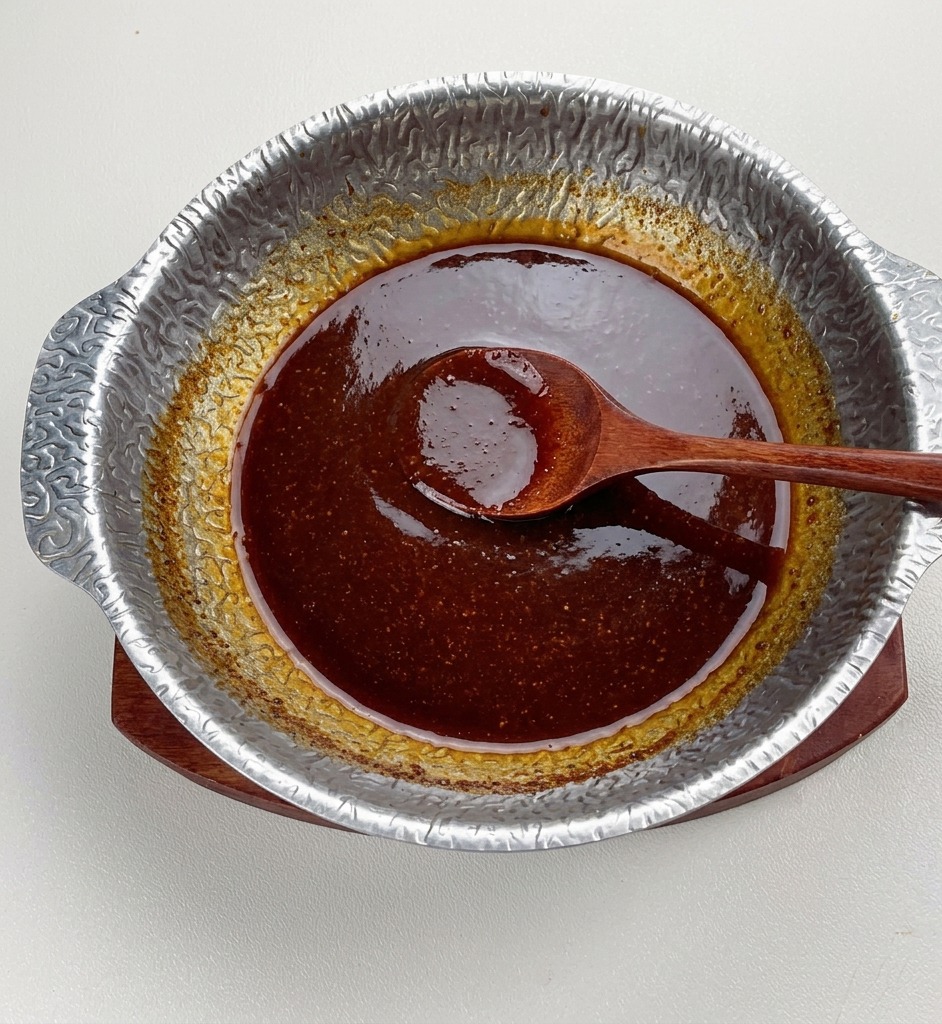 Die fertige dunkle Miso-Sauce köchelt in einem Metalltopf, sie sieht dickflüssig und glänzend aus, ein Holzlöffel ruht in der Mitte.