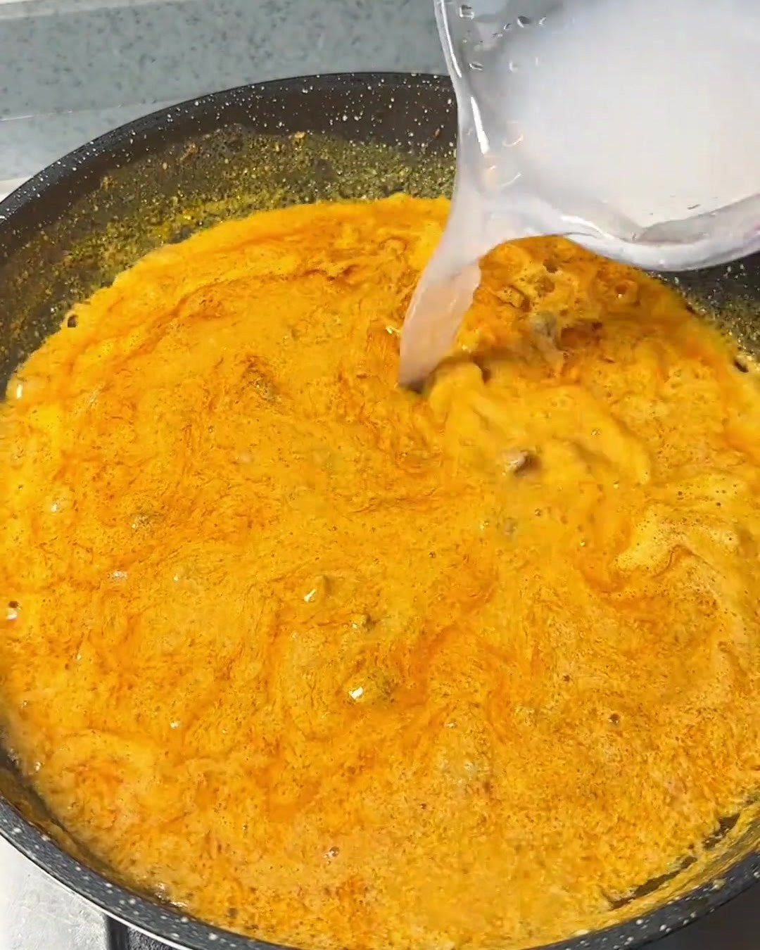 Klares Wasser wird aus einem Behälter in eine köchelnde, kräftig orangefarbene Curry-Mischung in einer dunklen Pfanne gegossen.