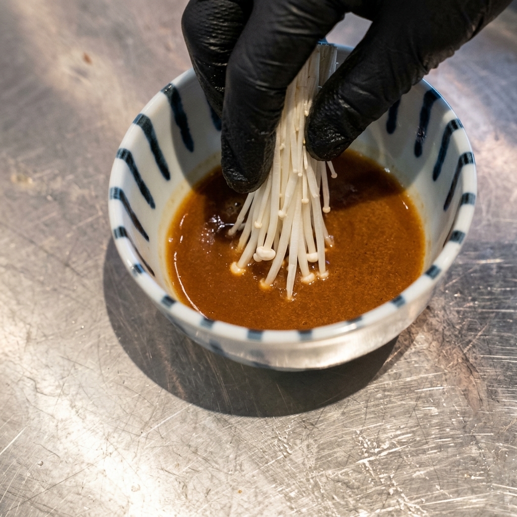 Eine Hand in einem schwarzen Handschuh legt eine Handvoll weiße Sprossen in eine Schale mit brauner Dip-Sauce.