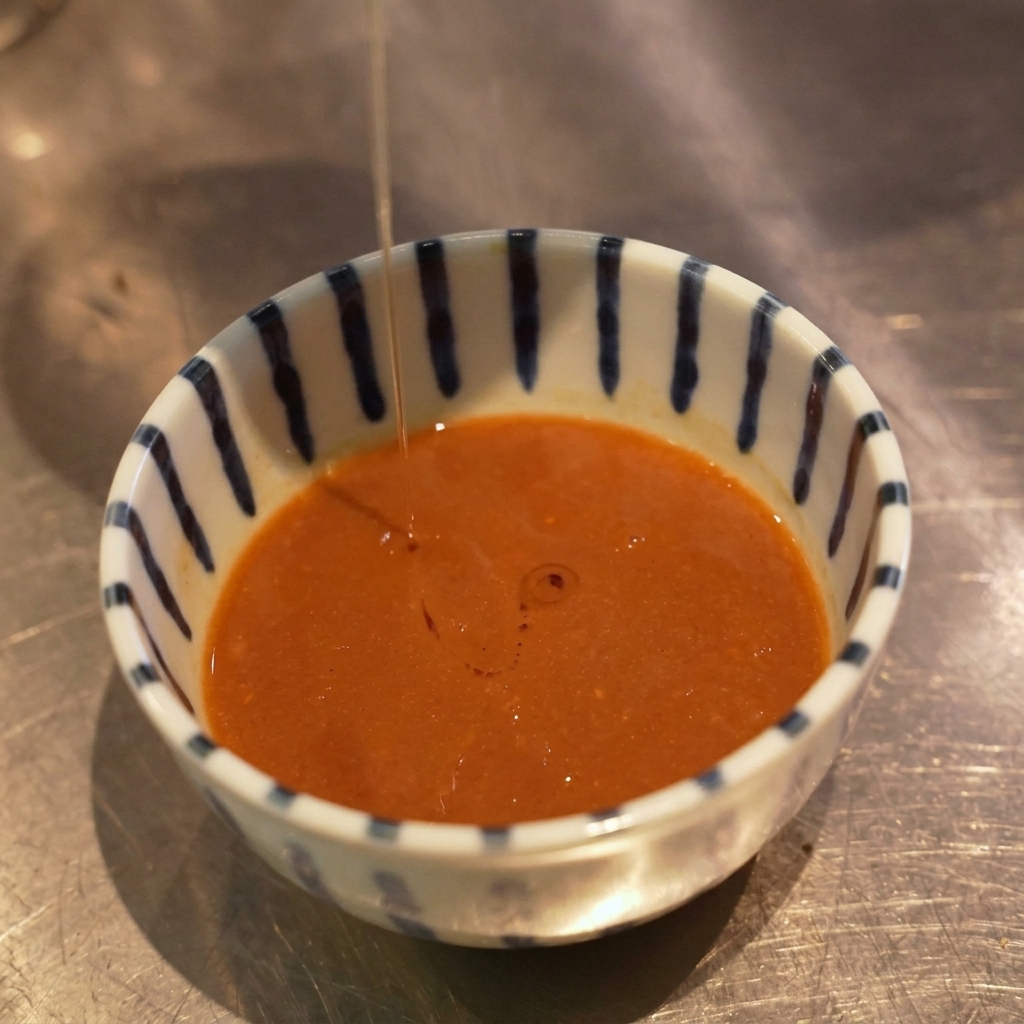 Ein dünner Strahl orangefarbenes Öl wird von oben in eine Schale mit Dip-Sauce geträufelt und bildet Tröpfchen auf der Oberfläche.
