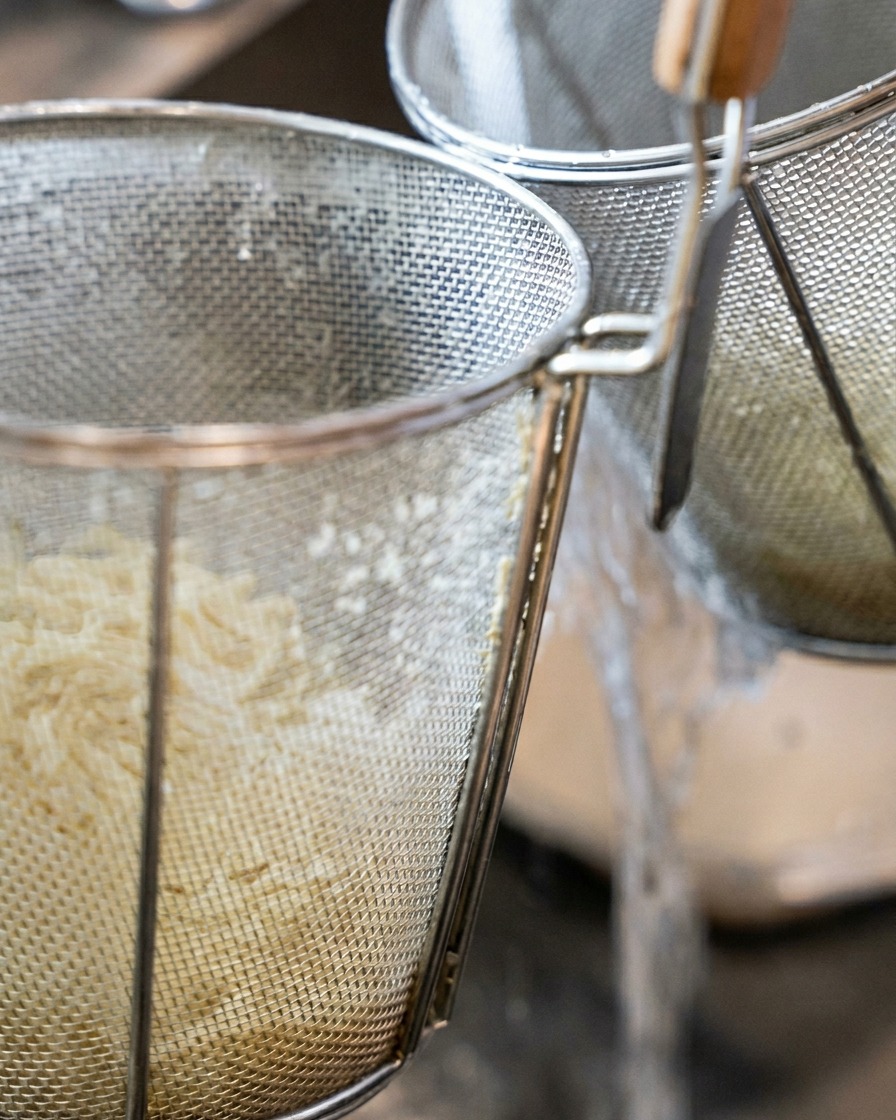 Dicke Ramen-Nudeln in einem zylindrischen Nudelsieb werden unter fließendem Wasser abgespült.