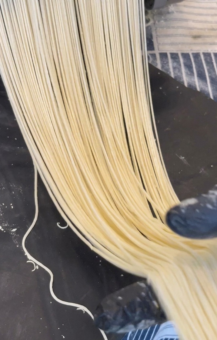 Lange Stränge hellgelber Ramen-Nudeln werden aus einer Metallmaschine auf eine dunkle Oberfläche gepresst.