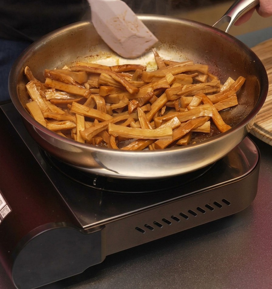 Ein weißer Spatel wendet die glasierten, braunen Bambussprossen in einer Bratpfanne, damit sie gleichmäßig mit dem duftenden Sesamöl überzogen werden.
