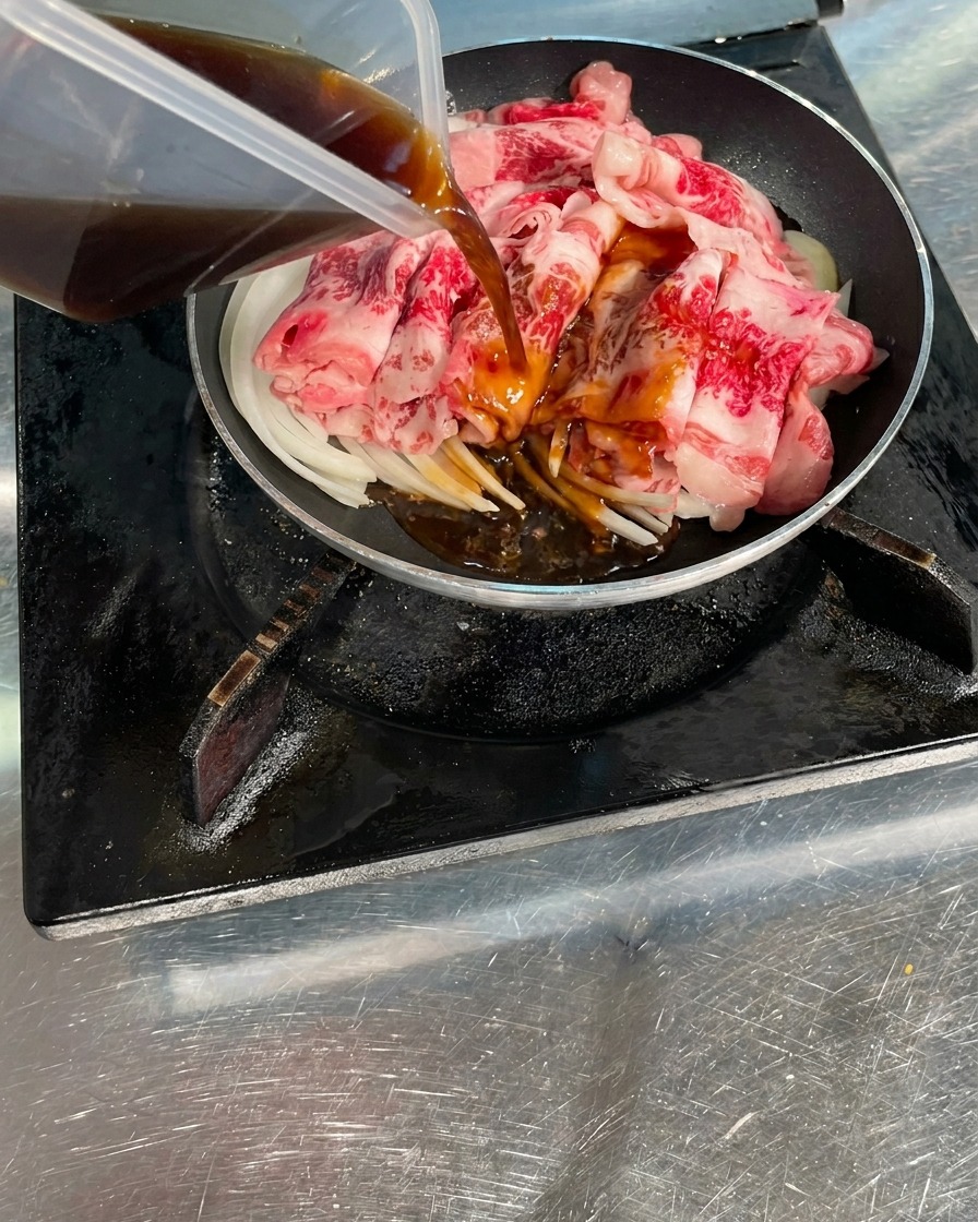 Dunkelbraune, herzhafte Sauce wird aus einem klaren Behälter über rohe Rindfleischscheiben und Zwiebeln in einer Kochpfanne gegossen.