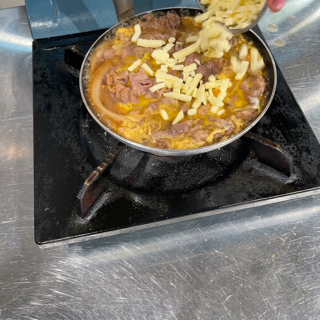 Eine Hand streut geriebenen weißen Käse über eine Pfanne mit gegartem Rindfleisch, Zwiebeln und leicht gestocktem Ei.