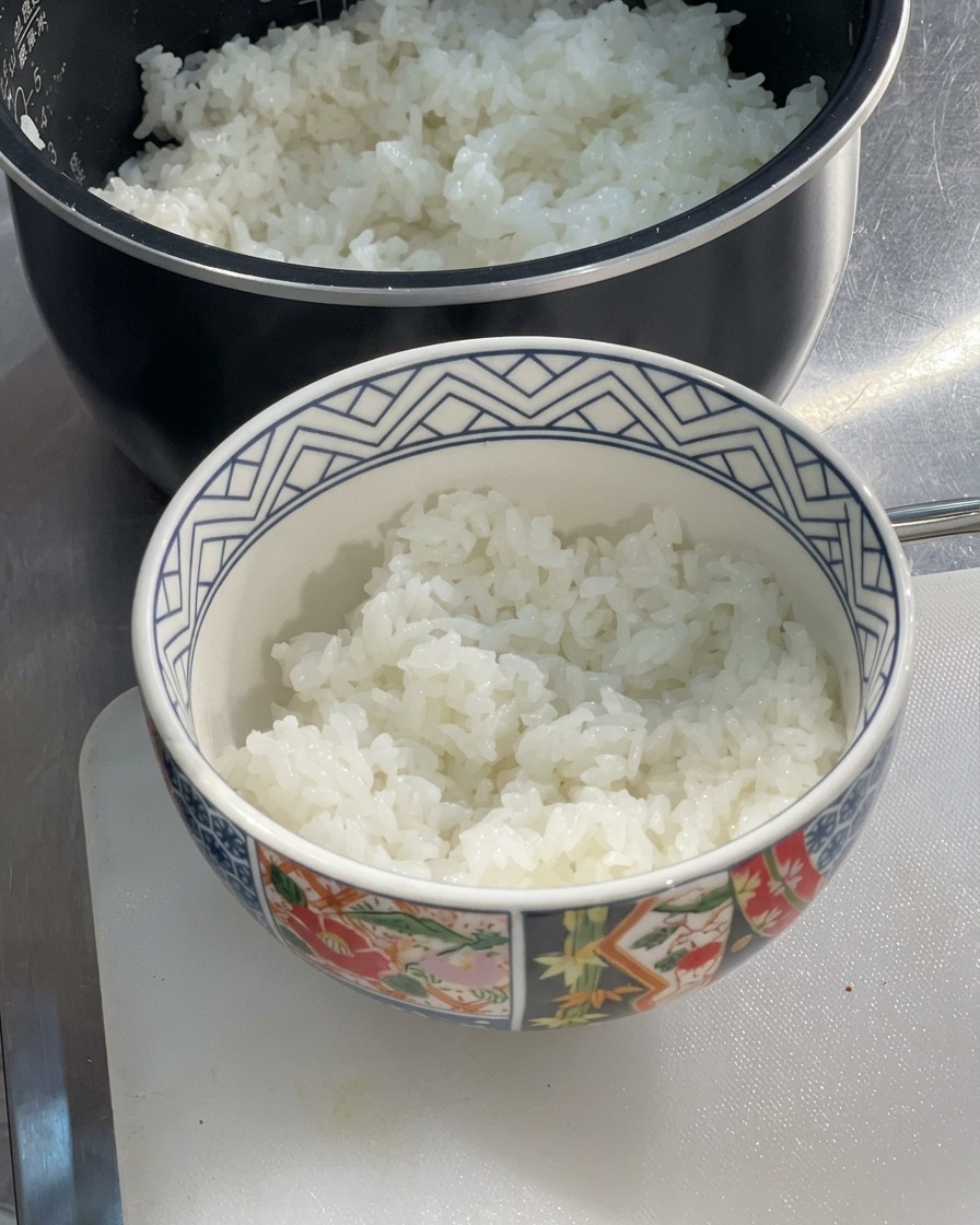 Eine gemusterte Keramikschale, halb gefüllt mit frisch gedämpftem, fluffigem weißem Reis, steht auf einem Schneidebrett neben einem Reiskocher.