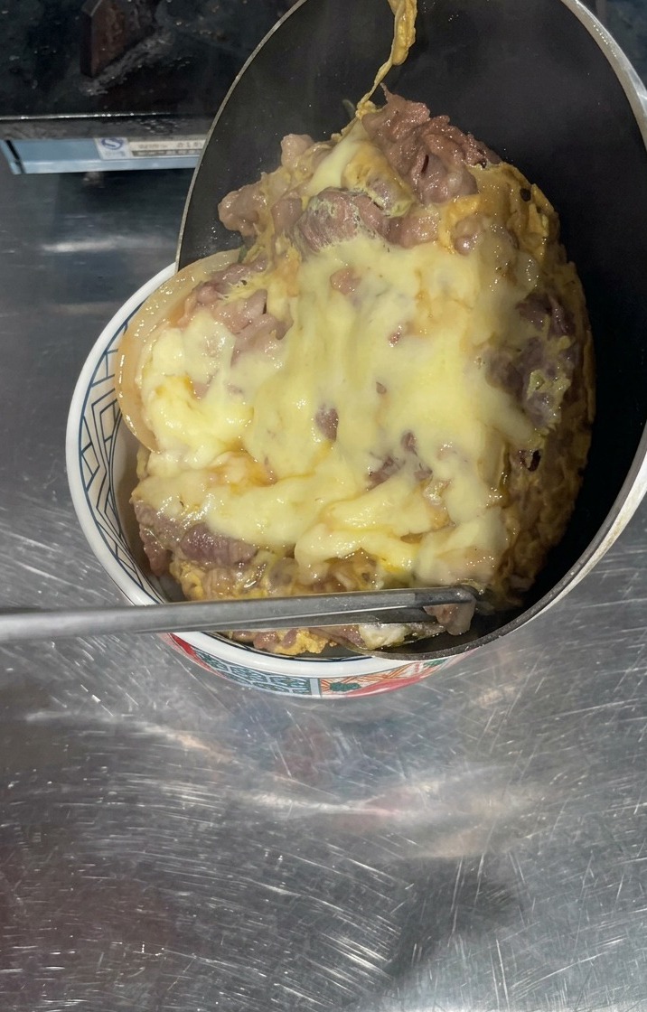 Eine Pfanne wird über eine Schüssel mit weißem Reis gekippt, wobei eine dicke Schicht aus gegartem Rindfleisch und geschmolzenem gelbem Käse herausgleitet, um die Oberfläche zu bedecken.
