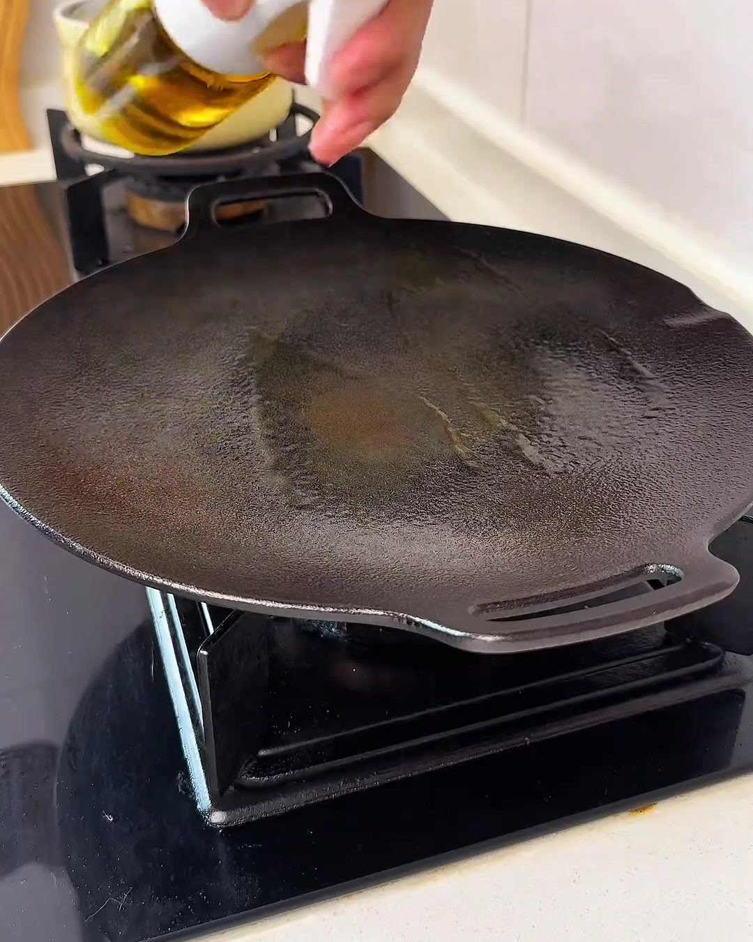 Eine Hand sprüht einen feinen Nebel aus goldenem Speiseöl auf eine saubere, schwarze runde Grillpfanne auf dem Herd.