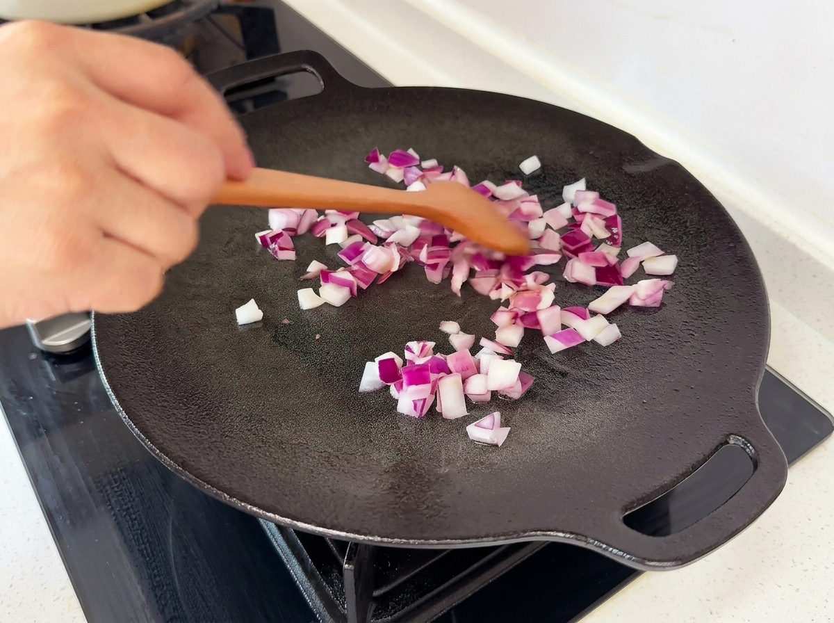 Gewürfelte rote Zwiebeln werden mit einem Holzspatel in einer schwarzen flachen Pfanne gewendet.