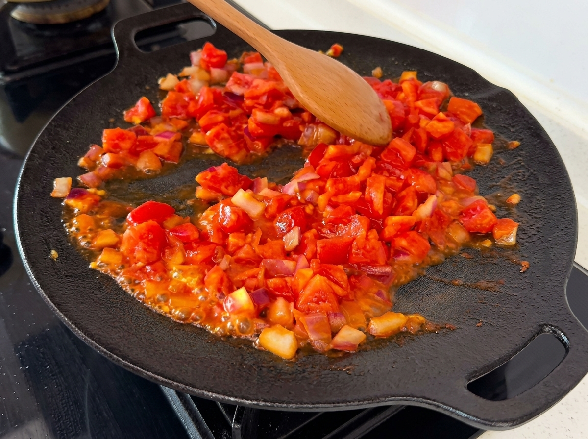 Zerkleinerte und gewürfelte Tomaten vermischen sich mit Zwiebeln in einer heißen Pfanne, wobei sichtbare Säfte brodeln.