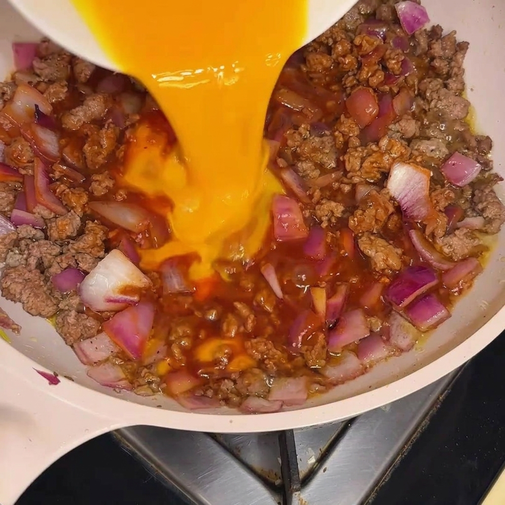 Die flüssige, gelbe Eimasse wird in einem gleichmäßigen Strom auf das köchelnde Fleisch-Zwiebel-Gemisch gegossen.