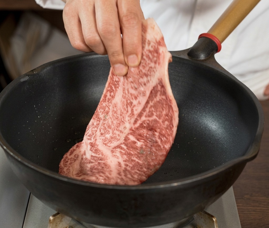 Eine Hand legt ein großes, gut marmoriertes und mit schwarzem Pfeffer gewürztes rohes Steak in eine heiße schwarze Pfanne.