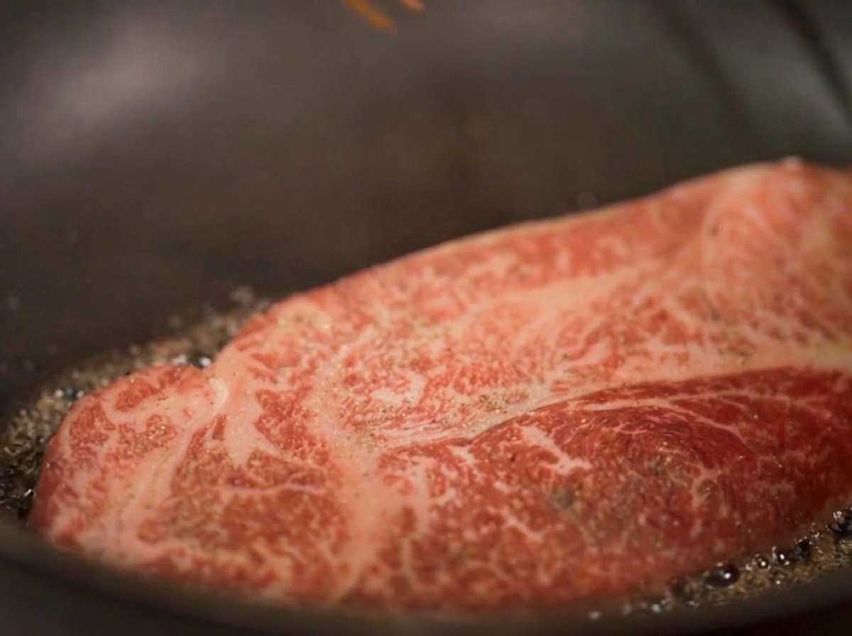 Nahaufnahme eines dicken Steaks, das in Öl brät, während Dampf aufsteigt und sich die unteren Ränder goldbraun färben.