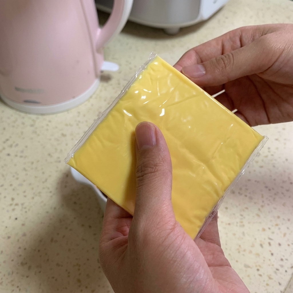 Eine Person hält zwei quadratische Scheiben hellgelben Schmelzkäses über eine Schüssel mit Rindfleisch und dunkler Sauce.