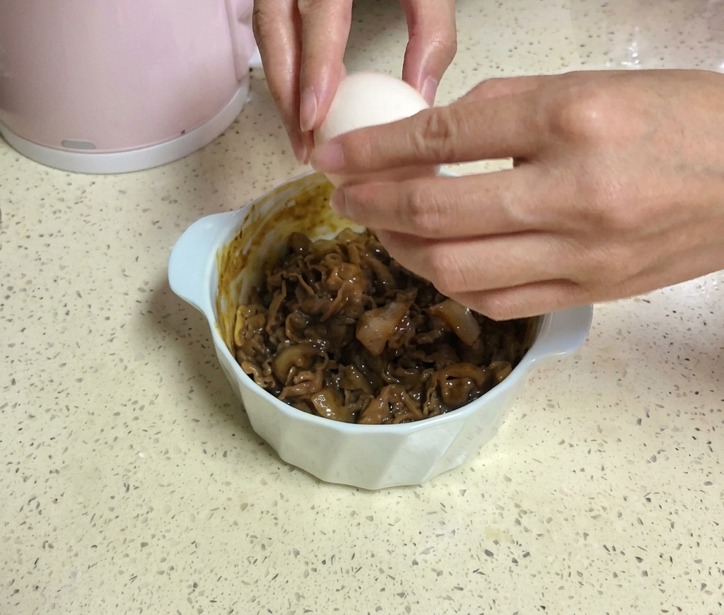 Zwei Hände schlagen ein weißes Ei über einer Schüssel mit Rindfleisch auf, die mit zwei Scheiben gelbem Käse belegt ist.