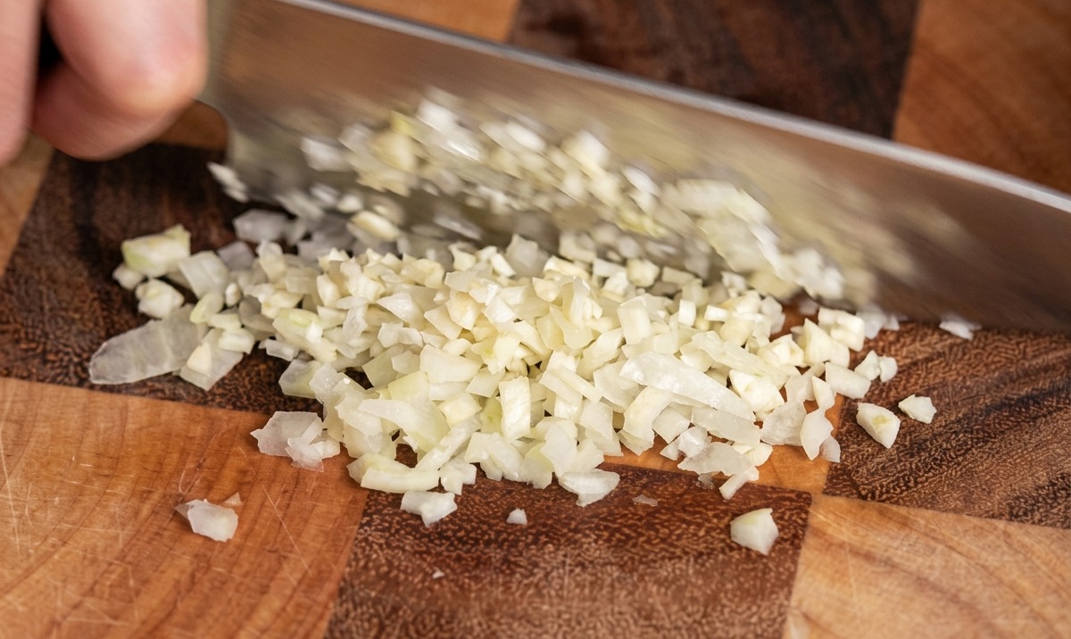 Nahaufnahme eines Kochmessers, das Knoblauchzehen auf einem Holzschneidebrett in feine Stücke hackt.