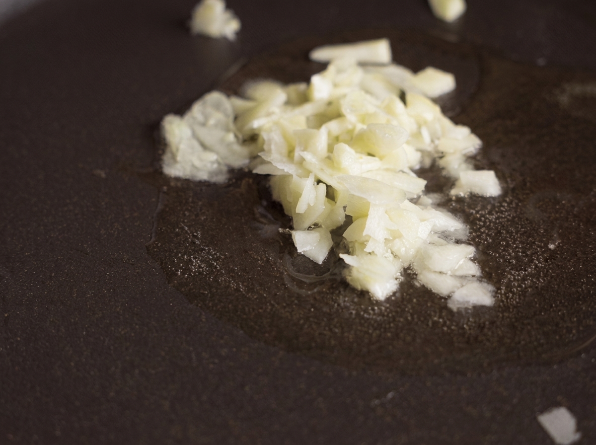 Fein gehackter Knoblauch brodelt in einer kleinen Menge klarem Öl in einer dunklen, beschichteten Bratpfanne.
