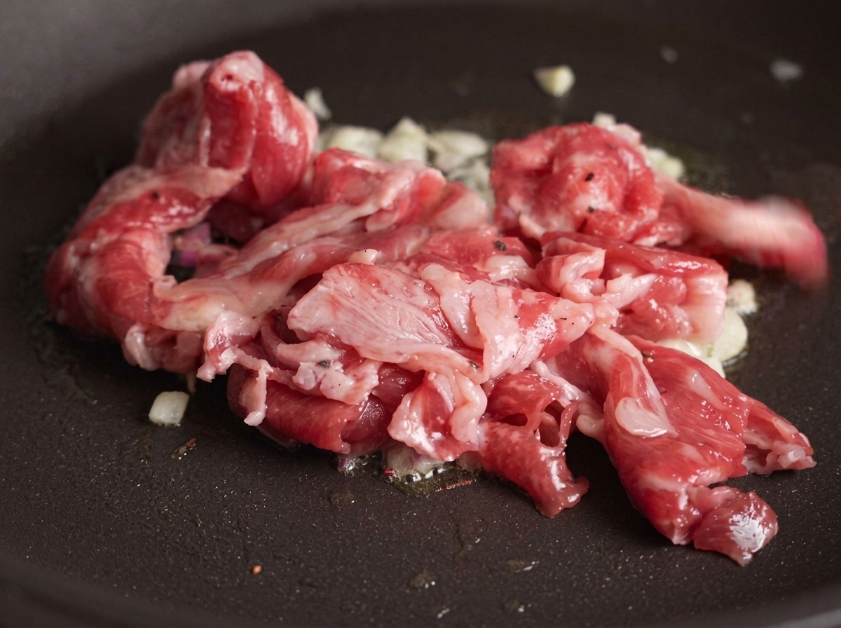 Rohe Rindfleischscheiben werden in die Pfanne zum angeschwitzten Knoblauch gegeben, wobei sich erste Stellen durch die Hitze braun färben.