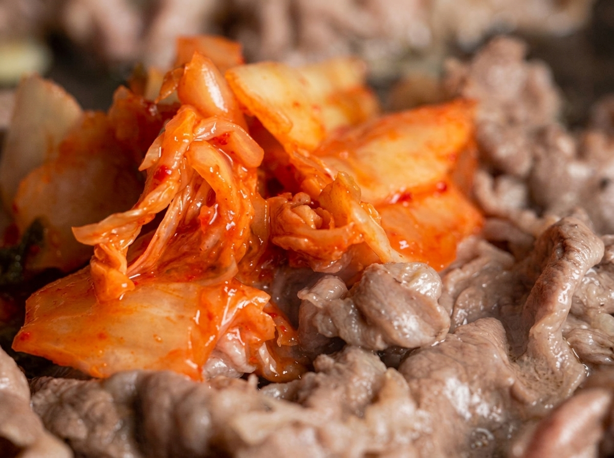 Eine großzügige Portion leuchtend rotes Kimchi wird in einer Bratpfanne unter die dünnen Rindfleischstreifen gemischt.