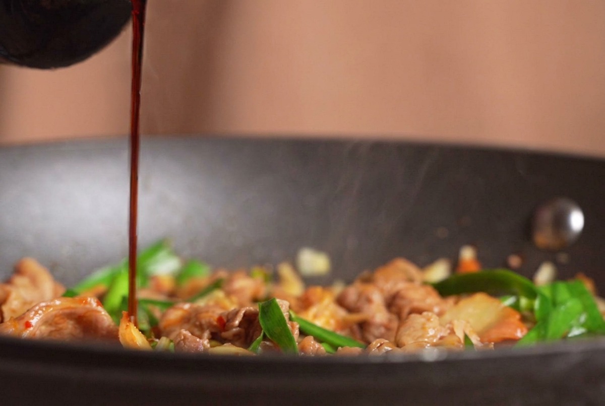Dunkle Sojasauce wird aus einer Flasche über die dampfende Pfanne mit Rindfleisch, Kimchi und Schnittknoblauch gegossen.